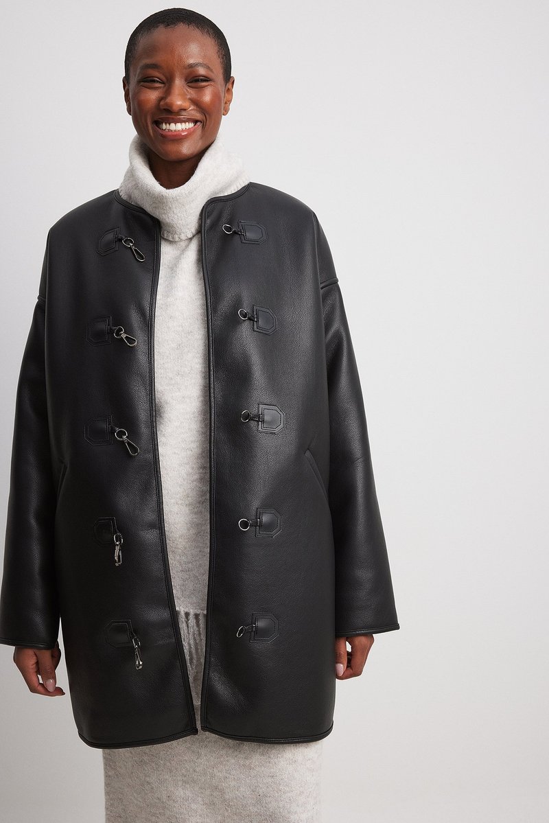 Juodas dirbtinės odos paltas su laisvu kirpimu, užsegamas metalinėmis sagtimis. Yra aukšta apykaklė ir lygus, matinio paviršiaus tekstūra.