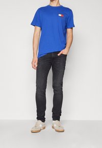 Royal blue t-shirt med en liten flerfärgad rektangulär logotyp, i kombination med mörka slim-fit jeans och ljusbeige sneakers med vita accenter.