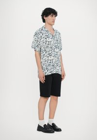 Λευκό πουκάμισο κοντομάνικο με μπλε φυτικό σχέδιο, συνδυασμένο με μαύρη βερμούδα. Το μοντέλο φοράει μαύρες λουστρίνι και λευκές κάλτσες.