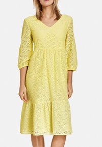 Vestido amarillo de encaje con escote en V, mangas tres cuartos y falda de volantes, que presenta un patrón de ojales para añadir textura.