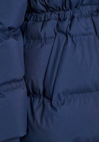 Tamsiai mėlynos pūkinės striukės detalė, parodanti pūkumo tekstūrą, elastingą juosmenį ir elegantišką šoninį kišenės dizainą su mažo juodo metalo akcentu.