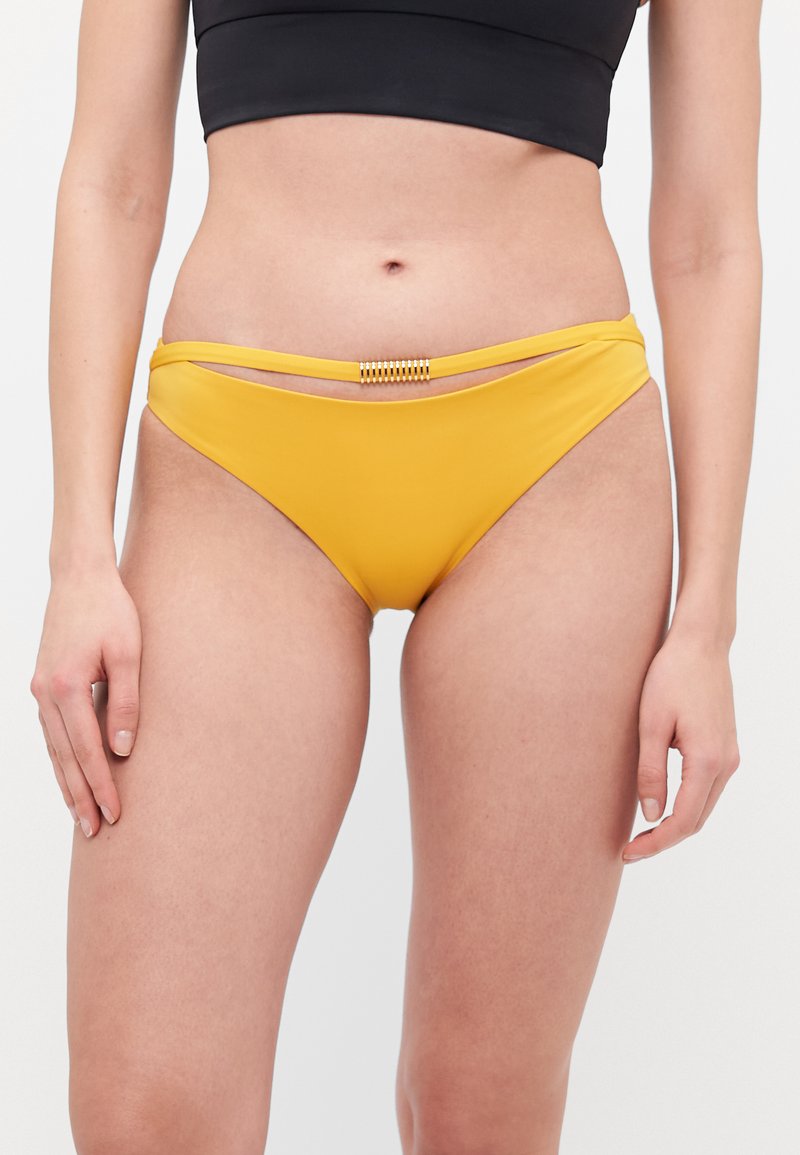 Gule bikini-bukser med glatt stoff, lav midje, og en dekorativ stropp med gullfarget beslag foran i midten.