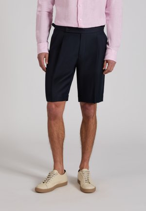 Uomo che indossa pantaloncini neri su misura, una camicia rosa a maniche lunghe e sneakers beige, in piedi davanti a uno sfondo semplice.