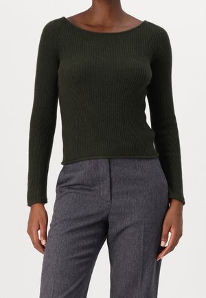 Person iført mørkegrøn ribstrikket langærmet top og grå bukser med fiskebenmønster, stående foran en ensfarvet baggrund.