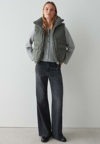 Grüne gepolsterte Weste mit hohem Kragen und zwei Vorderakttaschen über einem grauen gerippten Pullover, kombiniert mit schwarzen Schlagjeans und schwarzen Schuhen.