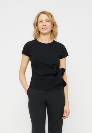 Blonde Frau trägt ein eng anliegendes schwarzes Kurzarmoberteil mit einer großen seitlichen Schleife, kombiniert mit schwarzen Hosen, steht vor einem schlichten hellen Hintergrund.