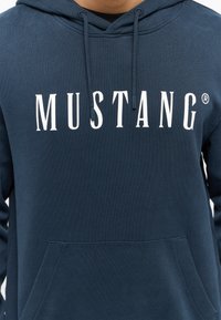 Felpa blu navy in misto cotone, con tasca a marsupio, cappuccio regolabile con cordino e grande logo bianco "MUSTANG®" stampato sul davanti.