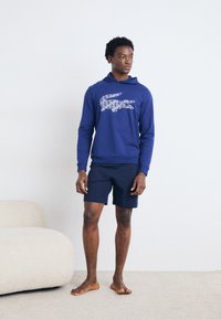 Herren Kapuzenpullover in Marineblau mit einem grafischen Krokodil-Design, aus weichem Material gefertigt und mit dunklen Shorts kombiniert. Lässige Passform, glatte Textur.