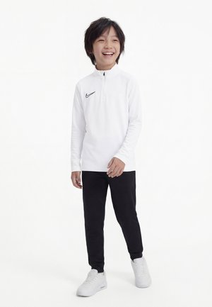 Enfant souriant portant un haut Nike blanc à manches longues, un pantalon noir et des baskets blanches, se tenant devant un fond blanc uni.