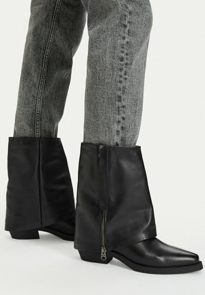 Black leather ankle boots with side zippers worn with faded gray jeans against a plain light background.
