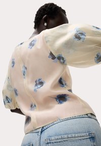 Blusa in crema trasparente con motivo floreale blu e maniche a sbuffo, abbinata a jeans azzurri. Include dettagli di cucitura delicati.