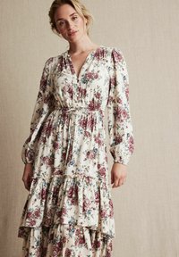 Kremowa sukienka maxi w kwiaty z fioletowymi i niebieskimi wzorami róż, z marszczoną talią, guzikami, długimi rękawami oraz falbanami na dole.