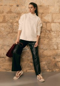 Witte sweatvest met ribbelkraag en reliëftekst, zwarte faux leren broek en beige-brouw sneakers. Houdt een bordeauxrood tasje vast.
