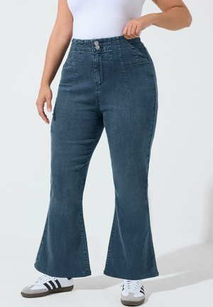 Kvinde iført mørkeblå jeans med høj talje og vide ben, kombineret med hvide sneakers og en hvid ærmeløs top, står med den ene hånd på hoften.