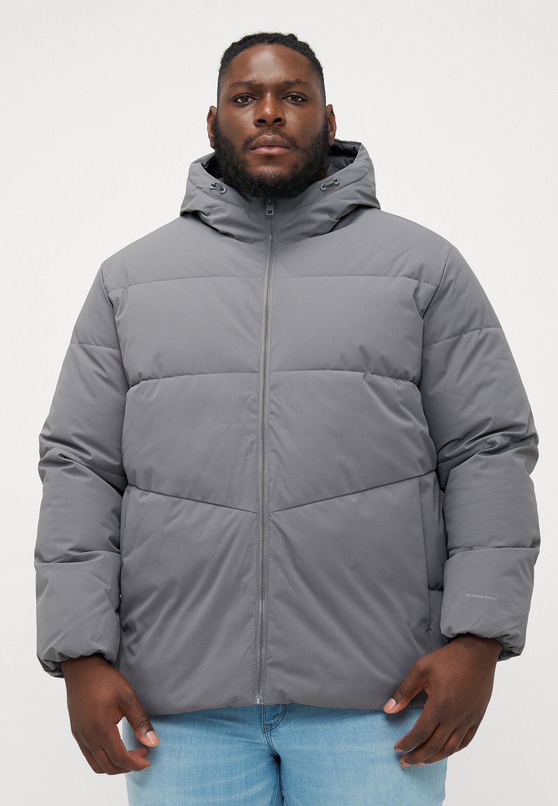 Chaqueta acolchada gris con capucha, que presenta un cierre con cremallera, un diseño acolchado y puños elásticos. El material parece suave con una textura lisa.