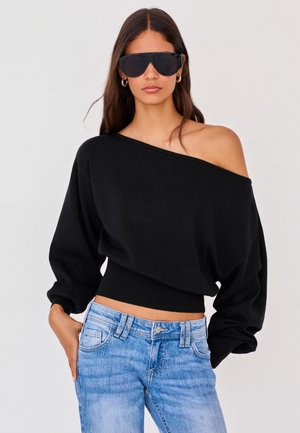 Kvinde iført oversized sort off-shoulder sweater, lyseblå jeans, store sorte solbriller og sølv runde øreringe, stående med den ene hånd i lommen.