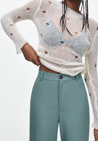 Maglione bianco in maglia con aperture, abbinato a pantaloni teal a vita alta con una texture liscia e dettaglio del bottone frontale.