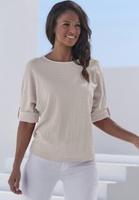 Beige strukturierte Bluse mit Rundhalsausschnitt und kurzen eingerollten Ärmeln, kombiniert mit weißen Hosen, vor einem hellen Hintergrund.