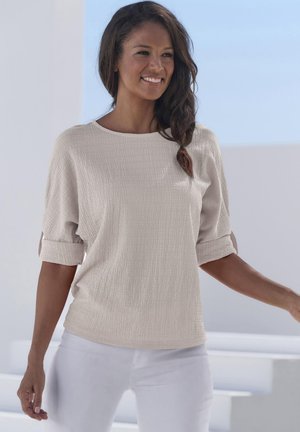 Beige strukturierte Bluse mit Rundhalsausschnitt und kurzen eingerollten Ärmeln, kombiniert mit weißen Hosen, vor einem hellen Hintergrund.