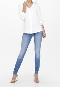 Blusa blanca de botones con escote en V y mangas largas; combinada con jeans ajustados azul claro y sandalias de tacón cuadrado transparentes.