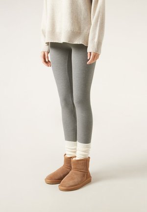 Persona que lleva pantalones ajustados grises acanalados, suéter crema, calentadores de piernas blancos y botines de ante marrón, de pie sobre un fondo claro.