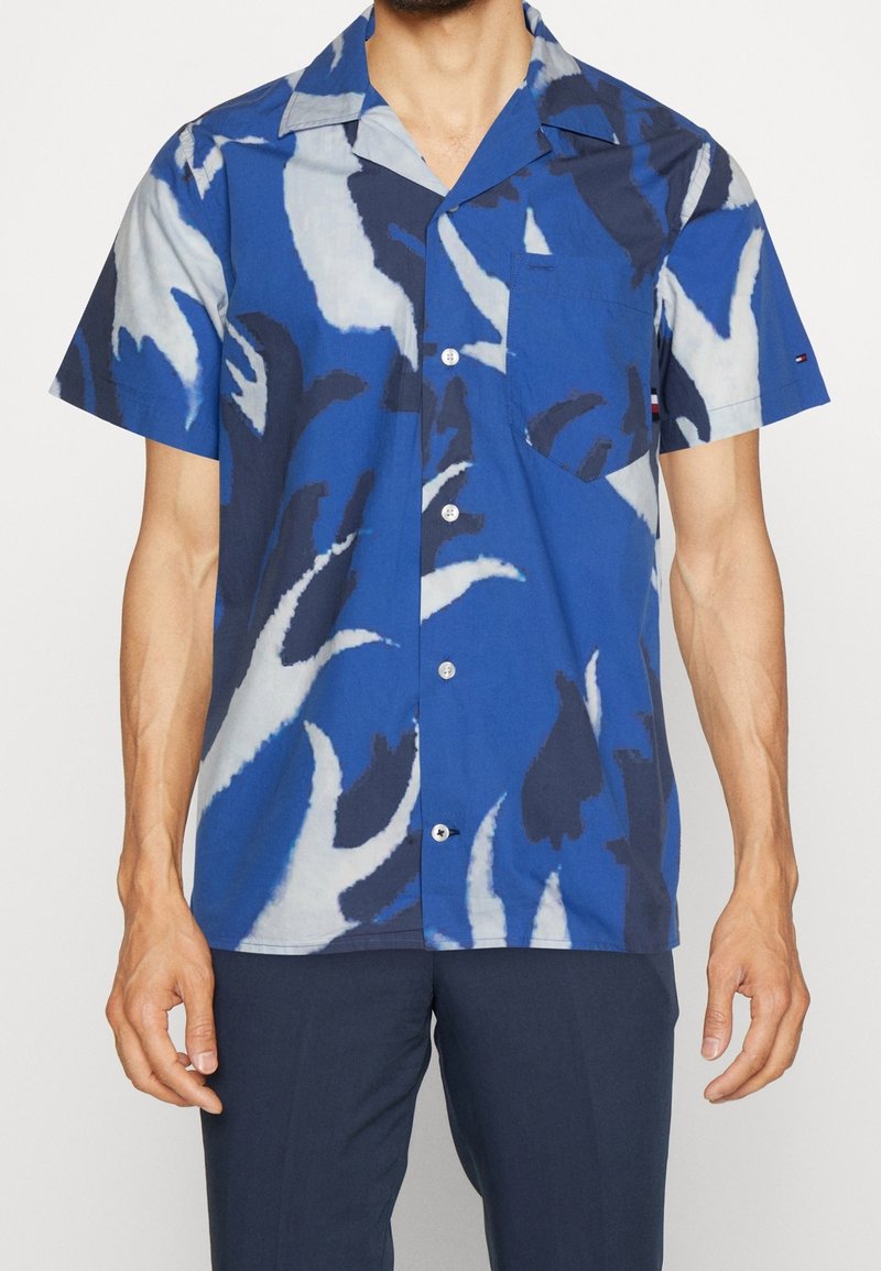 Camicia a maniche corte con bottoni di colore blu, decorata con motivi astratti bianchi e scuri, dotata di taschino sul petto e colletto a punte.