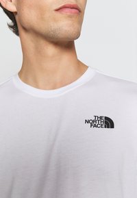 Camiseta de algodón blanca con cuello redondo y mangas cortas, que presenta un logo negro de "The North Face" en el lado izquierdo del pecho.
