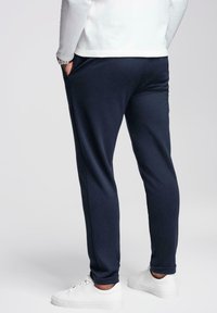 Pantalons slim-fit bleu marine portés par une personne debout les mains dans les poches, associés à des baskets blanches et un haut à manches longues blanc.