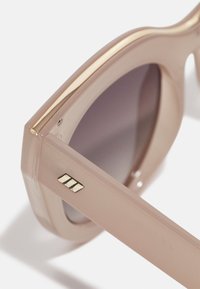 Sunglasses med en ljusrosa plastängel, guld-accenterad ovankant och tonade linser. Har en guldlogotyp på sidan av bygeln.