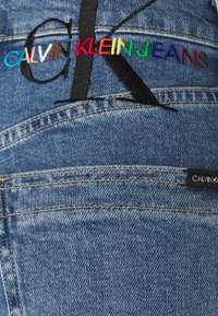 Denim fabric with embroidered "CALVIN KLEIN JEANS" logo in multicolored thread above a black embroidered "CK" and a small black Calvin Klein tag.