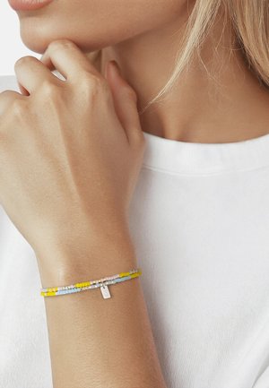 Primo piano di un polso con un delicato braccialetto di perline giallo, blu e rosa, indossato da una persona con un top bianco, mano vicino al mento.