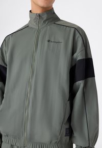 Mężczyzna w szarej i czarnej bluzie z zamkiem Champion z wysokim kołnierzem, elastycznym pasem i logo na lewej piersi oraz na dolnym boku.