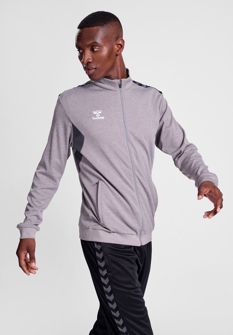 Grå zip-up atletisk jakke med sorte accenter og logo. Blødt stof, sidelommer, tætsiddende manchetter, parret med sorte træningsbukser med et mønstret stribe.