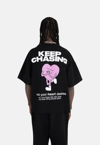 Svart oversized t-skjorte med en rosa hjertefigur og hvit tekst. Designet inkluderer en leken frase på baksiden.