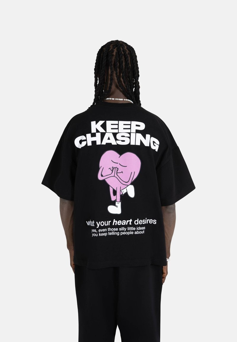 Svart oversized t-skjorte med en rosa hjertefigur og hvit tekst. Designet inkluderer en leken frase på baksiden.