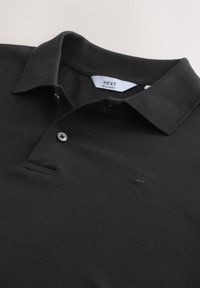 Mustast tekstuurkangast polo-särk, millel on lame krae, kahekordne nööbiliist ja rinnal väike reljeefne logo.