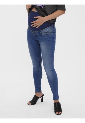 Gravid person iført blå ventetøj jeans og sorte højhælede sandaler løfter skjorten for at vise mavepanelet.