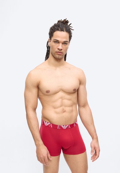 Μυώδης άνδρας με ντρέντλοκς που φοράει κόκκινα σχεδιαστικά μποξέρ με λευκή ζώνη μέσης με λογότυπο, στέκεται μπροστά σε απλό φωτεινό φόντο.