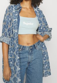 Niebieska koronkowa bluzka w kwiaty na jasnoniebieskim topie z napisem "Pegador". Zestawiona z dżinsami z wysokim stanem.