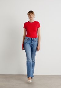T-shirt en coton rouge, jeans évasés bleu clair, tenant une pochette orange vif et des chaussures blanches sur fond uni.