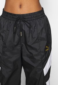 Svarta nylonjoggers med elastisk midja, dragsko och guld Puma-logotyp. Har vita sidopanels och fickor med blixtlås.