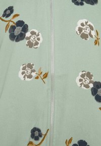 Tissu vert clair avec des imprimés floraux bleus et blancs et une fermeture éclair argentée qui court verticalement au centre.
