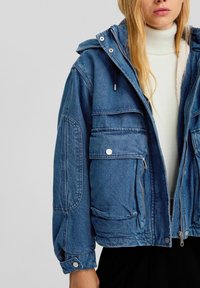 Femme portant une veste en denim bleue avec de grandes poches avant sur un pull à col roulé blanc et une jupe noire.