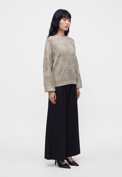 Femme aux longs cheveux noirs portant un pull beige à motifs, un large pantalon noir et des talons hauts pointus noirs, debout devant un fond uni.