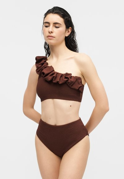 Femme portant un maillot de bain deux pièces marron texturé avec un haut à volants sur une épaule et un bas taille haute, debout devant un fond neutre.