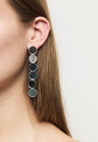 Ženské ucho ozdobené dlouhým náušnicí se šesti kulatými odrazovými diskem uspořádanými v vertikální linii na neutrálním pozadí.