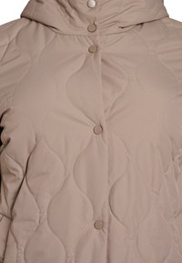 Tikkitakki, joka on beigen värinen ja siinä on huppu, nepparikiinnitys sekä aaltoileva kuvio. Kangas on sileäpintainen ja siinä on näkyvät ompelusaumat.