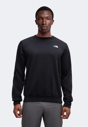 Homme portant un sweat-shirt noir à col rond avec le logo The North Face sur la poitrine et un pantalon gris, debout devant un fond uni.