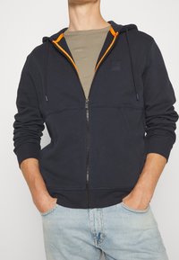 Sudadera con cremallera en color azul marino con acentos en naranja, bolsillos delanteros, puños acanalados y un acabado texturizado; usada sobre una camiseta verde apagada.