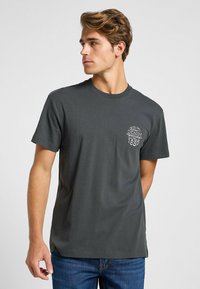 Camiseta de algodón gris oscuro con cuello redondo, mangas cortas y texto bordado en el lado izquierdo del pecho que dice "Denim de Calidad Desde 1889".
